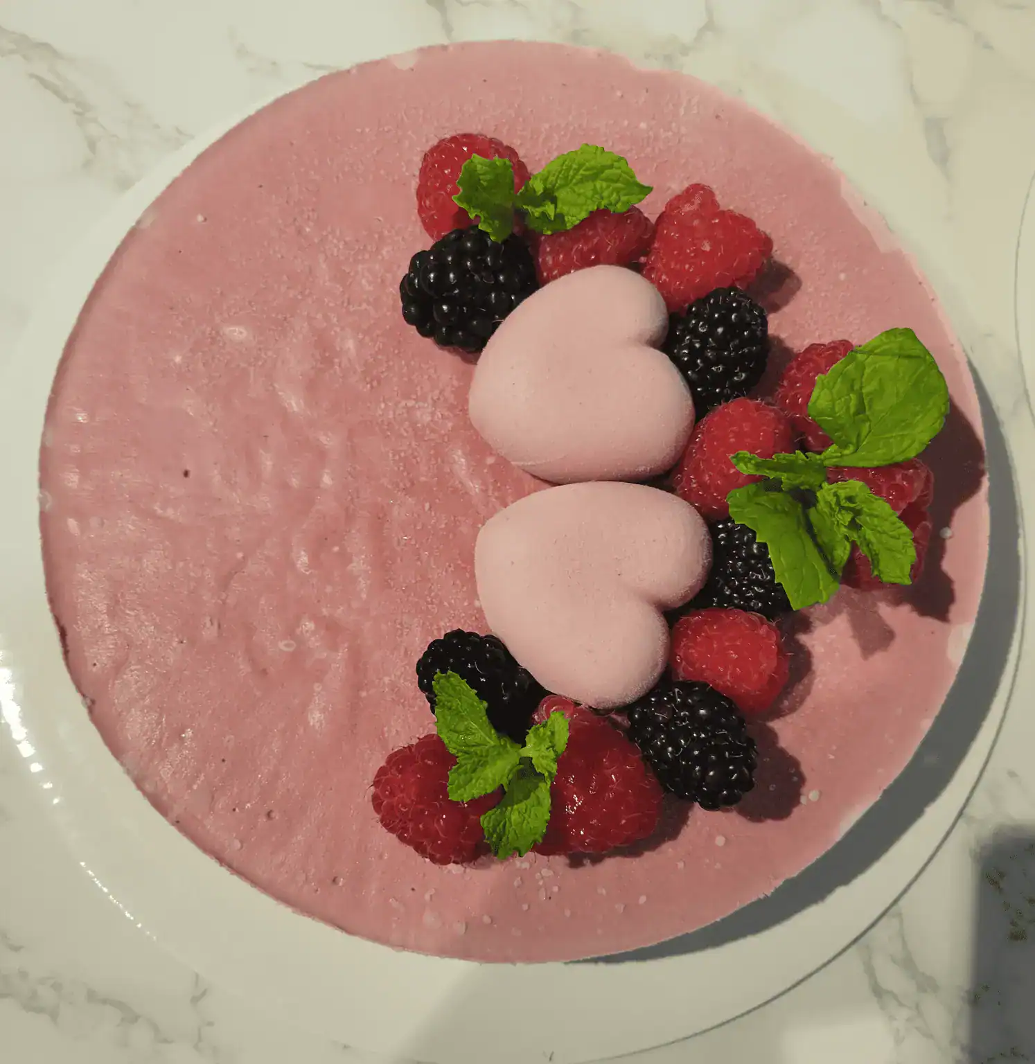 Strawberry Cheescake Raw vegan strawberry cheesecake topped with fresh raspberries, blackberries, mint leaves, and heart-shaped strawberry cream decorations on a marble surface.